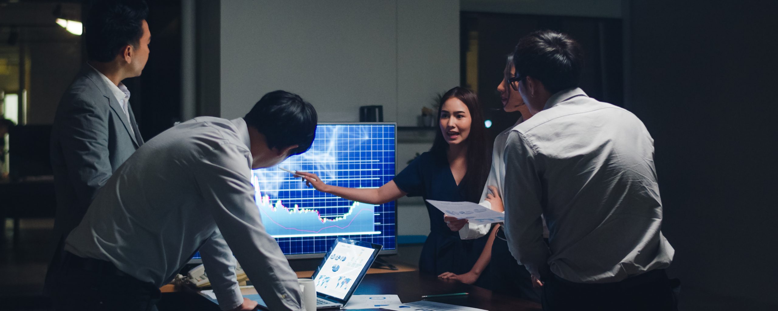Small group of professionals in an office meeting at night, gathered around a table with a laptop, printed charts and a large monitor