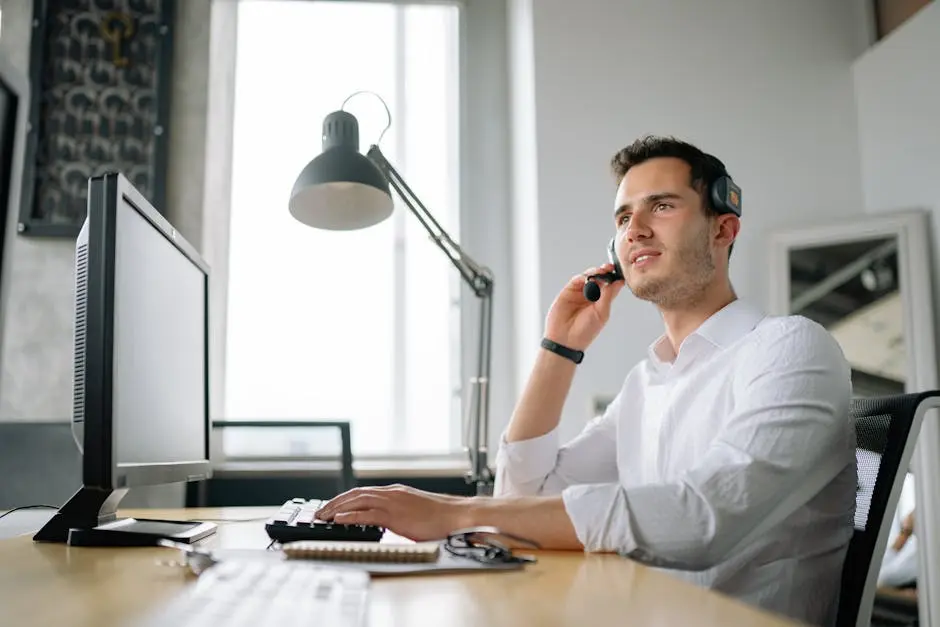 A professional young man wearing a headset and speaking into a microphone while seated at a desk with a desktop monitor, keyboard, notepad