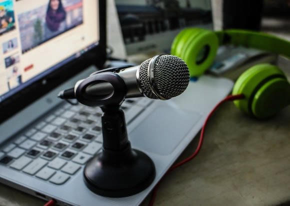 Close-up of a tabletop microphone on a stand placed on a laptop keyboard, with green over-ear headphones and a blurred computer screen in